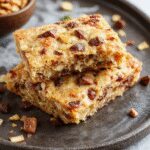 A close-up of vegan no-bake cookie dough bars arranged on a rustic wooden platter. The bars have a golden-brown exterior with visible specks of oat and chocolate chips, topped with a drizzle of dark chocolate and garnished with chopped nuts. The texture appears creamy yet firm, inviting and delicious.