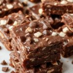 A close-up of a sliced brookie featuring a gooey chocolate brownie layer topped with a crisp cookie crust, showcasing its rich, fudgy interior and textured top, beautifully plated on a rustic wooden surface with some crumbs around.