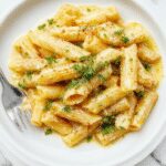 A close-up of a bowl of creamy penne pasta with visible garlic slices and a sprinkle of grated cheese on top. The pasta is coated in a smooth white sauce, garnished with fresh herbs, and served on a rustic plate that contrasts with the rich, velvety texture of the sauce.