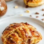 A golden, flaky croissant filled with rich, melted chocolate sits on a white plate. The pastry's layers are crispy and buttery, with a glossy finish from a light glaze. The chocolate oozes slightly from the open end, inviting and indulgent.