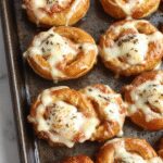 Golden-brown pretzels rellenos de jamón y queso, con una superficie ligeramente crujiente y exhibiendo un interior suave y fundido, presentados en un plato blanco sobre una mesa rústica, con algunos ingredientes visibles en el fondo.