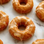 A close-up of three golden-brown baked pumpkin donuts drizzled with glossy maple glaze, arranged on a rustic white plate with a sprinkle of cinnamon. The donuts have a light, moist crumb with a slightly cracked surface, highlighting their fluffy texture. In the background, a small bowl of extra maple glaze and a few cinnamon sticks add warmth and seasonal charm.