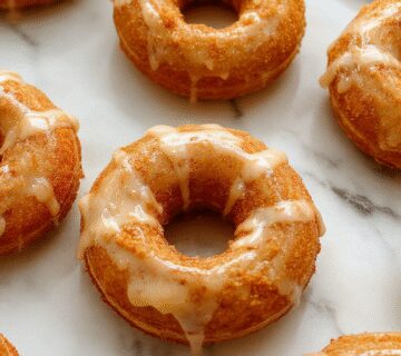A close-up of three golden-brown baked pumpkin donuts drizzled with glossy maple glaze, arranged on a rustic white plate with a sprinkle of cinnamon. The donuts have a light, moist crumb with a slightly cracked surface, highlighting their fluffy texture. In the background, a small bowl of extra maple glaze and a few cinnamon sticks add warmth and seasonal charm.
