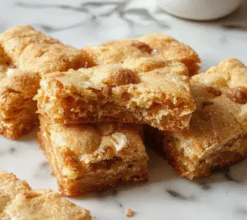 Un plato de blondies de remolino de canela en un molde rectangular, con un patrón de remolino visible en la superficie dorada y crujiente, adornados con un toque de canela espolvoreada y un pequeño racimo de canela en rama en el fondo, todo presentado sobre una mesa de madera rústica con utensilios de cocina y un fondo suave y luminosa.