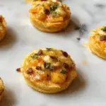 Mini cheesecake bites topped with a swirl of whipped cream and a sprinkle of autumn spices, displayed on a rustic wooden platter with scattered cinnamon sticks and pumpkin seeds, showcasing vibrant orange and creamy white colors with a textured surface and garnished with small pumpkin decorations.