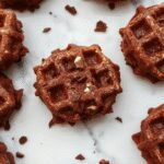 A golden-brown waffle topped with a glossy drizzle of chocolate sauce, sprinkled with powdered sugar, and garnished with fresh berries on a white plate. The waffle's crispy exterior contrasts with its soft interior, clearly showcasing its brownie-inspired texture. The presentation is inviting and elegant, with the background softly blurred to highlight the dessert.