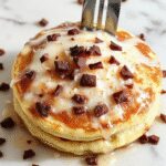 A stack of fluffy coconut cream pancakes topped with a generous dollop of coconut cream and garnished with shredded coconut and fresh berries on a white plate. The pancakes have golden edges and a soft, airy texture, with the coconut cream melting slightly over the top.
