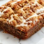 A rustic white plate holds a thick slice of moist banana bread with a golden-brown crust. The bread showcases a tender crumb dotted with banana chunks, with a slight glaze on top and a few banana slices as garnish. Soft natural lighting highlights the moist texture and inviting appearance, set against a neutral background for a cozy, homemade feel.