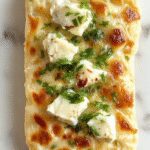Golden-brown cottage cheese flatbread cut into triangular slices, showcasing its soft interior and slightly crispy crust. The flatbread is topped with a sprinkle of herbs and served on a rustic wooden board, with a side of fresh greens and a drizzle of olive oil, inviting a wholesome and appetizing appearance.