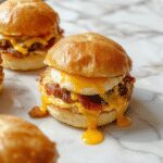 A close-up of three bacon egg and cheese sliders arranged on a white plate. Each slider has a toasted soft bun filled with crispy bacon, a fluffy scrambled egg, and melted cheese, topped with a small piece of bacon. The sliders are garnished with fresh herbs and appear steamed with the cheese slightly melted, showcasing a hearty and inviting breakfast presentation.