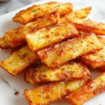 A close-up of golden-brown, crispy air fryer apple fries arranged on a rustic white plate, sprinkled with cinnamon sugar, with a few apple slices visible in the background, highlighting their crunchy texture and appealing color