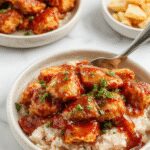 A bubbling crockpot filled with tender, glazed chicken pieces coated in a shiny, caramelized teriyaki sauce. The chicken is topped with sesame seeds and sliced green onions, with a side of steamed rice and colorful vegetables in the background, creating an inviting and appetizing scene.