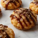 A close-up of a plate of golden-brown pumpkin syrup cookies with a glossy glaze, garnished with a sprinkle of cinnamon and chopped walnuts. The cookies are arranged on a rustic wooden surface with a few scattered pecans nearby, showcasing their soft and chewy texture with slightly cracked tops and melted chocolate chips.