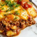 A hearty serving of Hobo Casserole on a rustic plate showing layers of golden browned ground beef, sliced potatoes, and melted cheese topping, garnished with fresh herbs, all presented on a wooden table with a spoonful served revealing its comforting, cheesy interior.