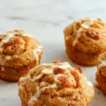 Golden-brown muffins topped with a swirl of cinnamon and pumpkin filling, arranged on a rustic wooden platter with a background of autumn leaves and cinnamon sticks. The muffins' crumb looks moist and tender, with visible specks of pumpkin and cinnamon swirl throughout.