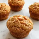 A close-up of a freshly baked muffin topped with dark chocolate chips, golden-brown with a slightly cracked surface, in a rustic paper wrapper, set on a wooden surface with a few scattered chocolate chips and a hint of cinnamon in the background.
