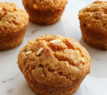 A close-up of a freshly baked muffin topped with dark chocolate chips, golden-brown with a slightly cracked surface, in a rustic paper wrapper, set on a wooden surface with a few scattered chocolate chips and a hint of cinnamon in the background.