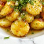 A close-up of golden-brown roasted potatoes with caramelized onion slices on top, served in a rustic ceramic dish. The potatoes are crispy on the outside with a soft interior, garnished with fresh herbs, and presented against a warm, inviting background that emphasizes comfort food.