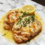 Un plato de pollo piccata con jugoso pollo dorado cubierto con una salsa brillante de limón, decorado con rodajas de limón y perejil fresco, presentado en un plato blanco elegante con una salsa ligeramente espesa y glaseada.