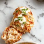 Un plato de pollo cremoso con una salsa blanca espesa y suave, cubierto con especias y trozos de pollo jugoso en un recipiente de cerámica rústica. Se acompaña con ramitas de perejil fresco y una cucharada de salsa ranch, todo presentado sobre una mesa de madera natural con utensilios de cocina a la vista.