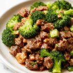 A vibrant stir-fry featuring tender ground beef glazed with a glossy honey garlic sauce, nestled among bright green broccoli florets on a white plate. The dish is garnished with a sprinkle of sesame seeds and chopped green onions, with a sautéed garlic clove visible at the edge, all set against a rustic wooden table.