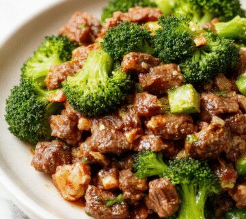 A vibrant stir-fry featuring tender ground beef glazed with a glossy honey garlic sauce, nestled among bright green broccoli florets on a white plate. The dish is garnished with a sprinkle of sesame seeds and chopped green onions, with a sautéed garlic clove visible at the edge, all set against a rustic wooden table.