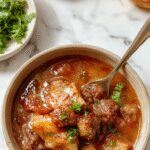 A close-up of a rich, hearty ragú de res served in a rustic bowl. The stew features tender chunks of beef coated in a glossy, savory tomato-based sauce, garnished with fresh herbs and accompanied by a side of crusty bread. The background showcases a wooden table with steam rising from the warm dish, highlighting its comforting appeal.