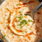 Cremoso dip de pollo buffalo en una taza blanca, adornado con gotas de salsa buffalo brillante y queso derretido, acompañado de palitos de apio y zanahoria al lado, todo sobre una mesa de madera rústica.