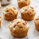 Un plato de muffins de pera especiada apilados en un plato blanco sobre una mesa de madera, con algunas peras frescas y especias como canela y nuez moscada dispersas alrededor. La corteza dorada y la textura esponjosa de los muffins son claramente visibles, con pedazos de pera en su interior. La presentación es acogedora y otoñal, con un fondo suave y cálido que realza los tonos cálidos de los muffins y las peras.