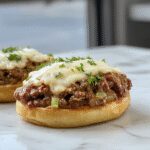 Un sándwich Sloppy Joe en un plato blanco con pan suave, carne molida cubierto con cebolla francesa caramelizada y queso fundido, acompañado de papas fritas y una ensalada fresca, todo en un ambiente informal y acogedor.