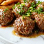 A close-up of tender Salisbury steak meatballs arranged neatly on a rustic ceramic plate, coated with rich brown gravy, garnished with fresh chopped parsley, and accompanied by mashed potatoes and steamed vegetables in the background.