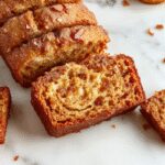 A golden-brown loaf of snickerdoodle banana bread sits on a rustic wooden surface, topped with a cinnamon-sugar crust. The bread's surface is slightly cracked, revealing a moist interior, with cinnamon-sugar sprinkled on top for added texture. A few slices are cut, showcasing a tender crumb speckled with hints of banana and cinnamon.