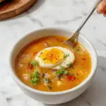 A steaming bowl of Sopa de Huevo Caído, featuring a clear golden broth with delicate, soft egg ribbons floating on top, garnished with chopped green onions and a few slices of soft tofu. The bowl is placed on a rustic wooden table, with a spoon partially submerged, capturing the inviting steam rising from the hot soup. The vibrant yellow of the eggs contrasts beautifully with the clear broth, creating a comforting and appetizing visual.