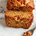 A slice of Starbucks copycat banana bread on a rustic white plate, showcasing its moist golden crumb with visible banana chunks and a slightly cracked top, served alongside a cup of steaming coffee on a wooden table.