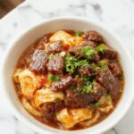 A vibrant bowl of sticky beef noodles featuring tender slices of beef atop glossy, caramel-colored noodles garnished with chopped green onions and sesame seeds. The dish is presented in a rustic white bowl on a wooden table, with steam gently rising, highlighting the luscious sauce coating the noodles and meat.