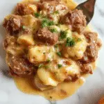 A close-up of a creamy beef stroganoff served over a bed of rice or noodles in a white, shallow dish. The beef is cooked in a rich, brown sauce with visible chunks of ground beef, garnished with fresh chopped parsley. The dish has a glossy, velvety texture with a slight sheen, and the sauce is thick and smooth, contrasting with the tender meat pieces and the fluffy rice or pasta. The background is softly blurred, highlighting the appetizing presentation and vibrant colors of the dish.