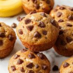 A close-up of two golden-brown banana chocolate chip muffins on a rustic wooden surface, with visible chocolate chips melting slightly on top. The muffins have a moist texture with a slightly cracked surface, and a few banana slices and chocolate chips are visible inside the crumb. Soft natural lighting highlights the moist interior and inviting aroma, with a slight sheen on the surface indicating freshness.