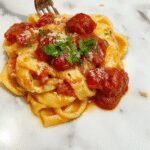 A close-up of a bowl of creamy tomato pasta topped with fresh basil leaves. The pasta has a rich, velvety crimson sauce with specks of herbs and grated cheese, garnished with a drizzle of olive oil and a sprinkle of black pepper. The dish is presented on a rustic wooden table with a sprig of basil beside it, evoking a warm and inviting atmosphere.