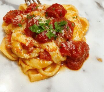 A close-up of a bowl of creamy tomato pasta topped with fresh basil leaves. The pasta has a rich, velvety crimson sauce with specks of herbs and grated cheese, garnished with a drizzle of olive oil and a sprinkle of black pepper. The dish is presented on a rustic wooden table with a sprig of basil beside it, evoking a warm and inviting atmosphere.