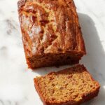A golden-brown zucchini bread loaf sliced to reveal its moist interior, with finely grated zucchini visible. The bread rests on a rustic wooden surface, with a few crumbs scattered around, and the texture looks soft and tender with a slightly crispy crust.