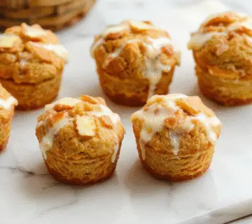 A close-up image of freshly baked Greek yogurt apple cinnamon muffins arranged on a rustic wooden platter. The muffins have a golden-brown top with a slightly cracked surface, speckled with tiny bits of apple and cinnamon. One muffin is broken open showing its moist, fluffy interior dotted with bits of apple and a swirl of cinnamon, while a few fresh apple slices are placed beside the muffins for garnish. The background is softly blurred, emphasizing the textures and warm tones of the baked goods.