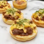 A rustic white plate featuring tender chunks of beef stew with golden-brown edges, topped with melted butter and finely chopped garlic, surrounded by crispy roasted potatoes. The dish is garnished with fresh herbs and served with a side of crusty bread. The presentation emphasizes the savory, hearty nature of the stew with warm, inviting colors and textures.