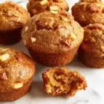 A close-up of freshly baked apple cinnamon muffins arranged on a rustic wooden platter, golden-brown tops sprinkled with cinnamon sugar, with visible chunks of apple and swirls of cinnamon inside. The muffins are showcased against a soft-focus background featuring autumn leaves and a cozy kitchen setting.